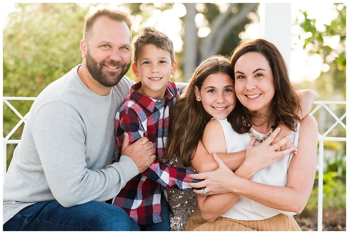 A smiling family of four sitting outdoors, with green foliage in the background. The father has a beard and is wearing a light gray sweater. The mother has shoulder-length brown hair and is wearing a white top. The daughter has long brown hair and is
