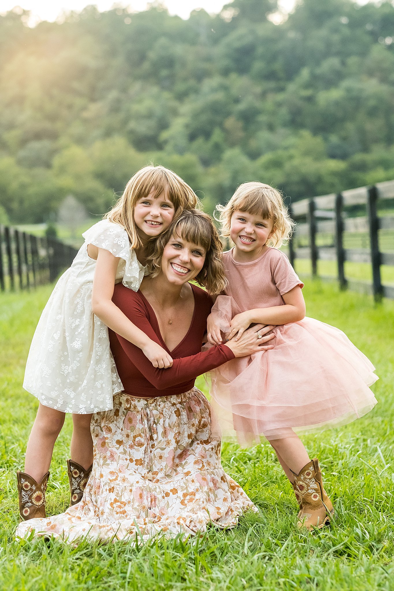 A woman kneeling on the grass with two young girls, all smiling and looking at the camera, outdoors in a grassy field with a fence and trees in the background.