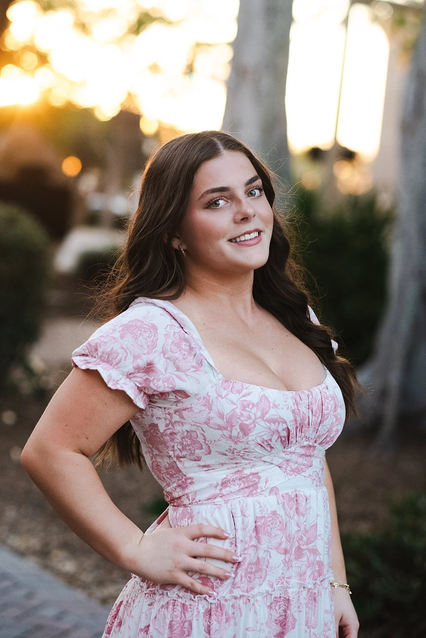 A young woman with long brown hair, wearing a pink and white floral dress, is outdoors during sunset, standing with one hand on her hip and smiling at the camera.
