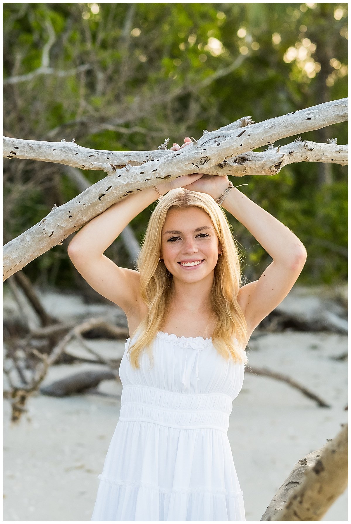 A young woman with long blonde hair smiling at the camera, holding a large tree branch above her head on a beach with green trees in the background.