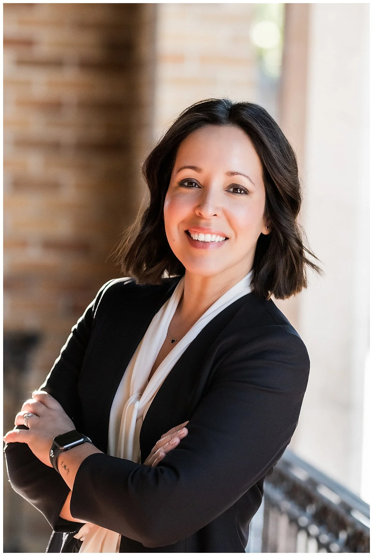 A woman with shoulder-length dark hair smiling, wearing a black blazer over a white blouse, standing with arms crossed in front of a window and brick wall.