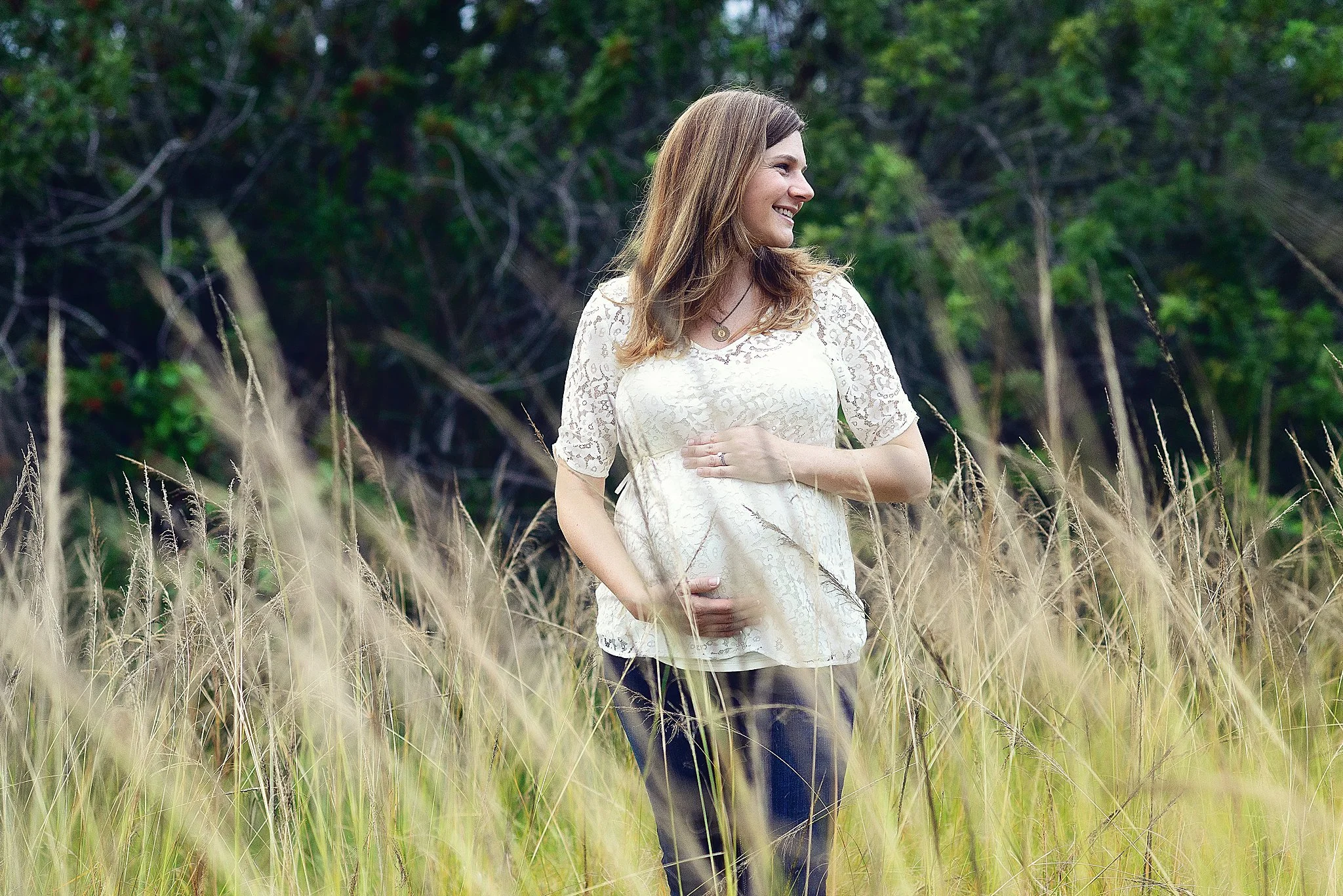 A smiling woman in a white lace top and dark pants standing in a grassy field with trees in the background.