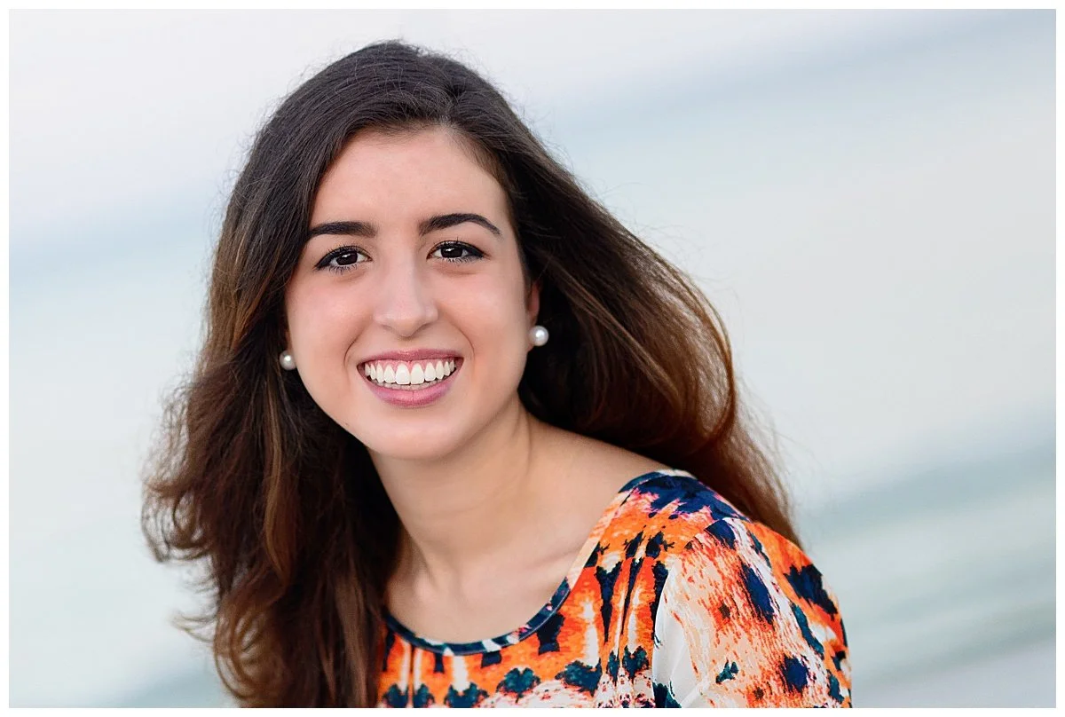 A young woman with long brown hair, smiling at the camera, wearing pearl earrings and a colorful patterned top, outdoors near water with a cloudy sky in the background.