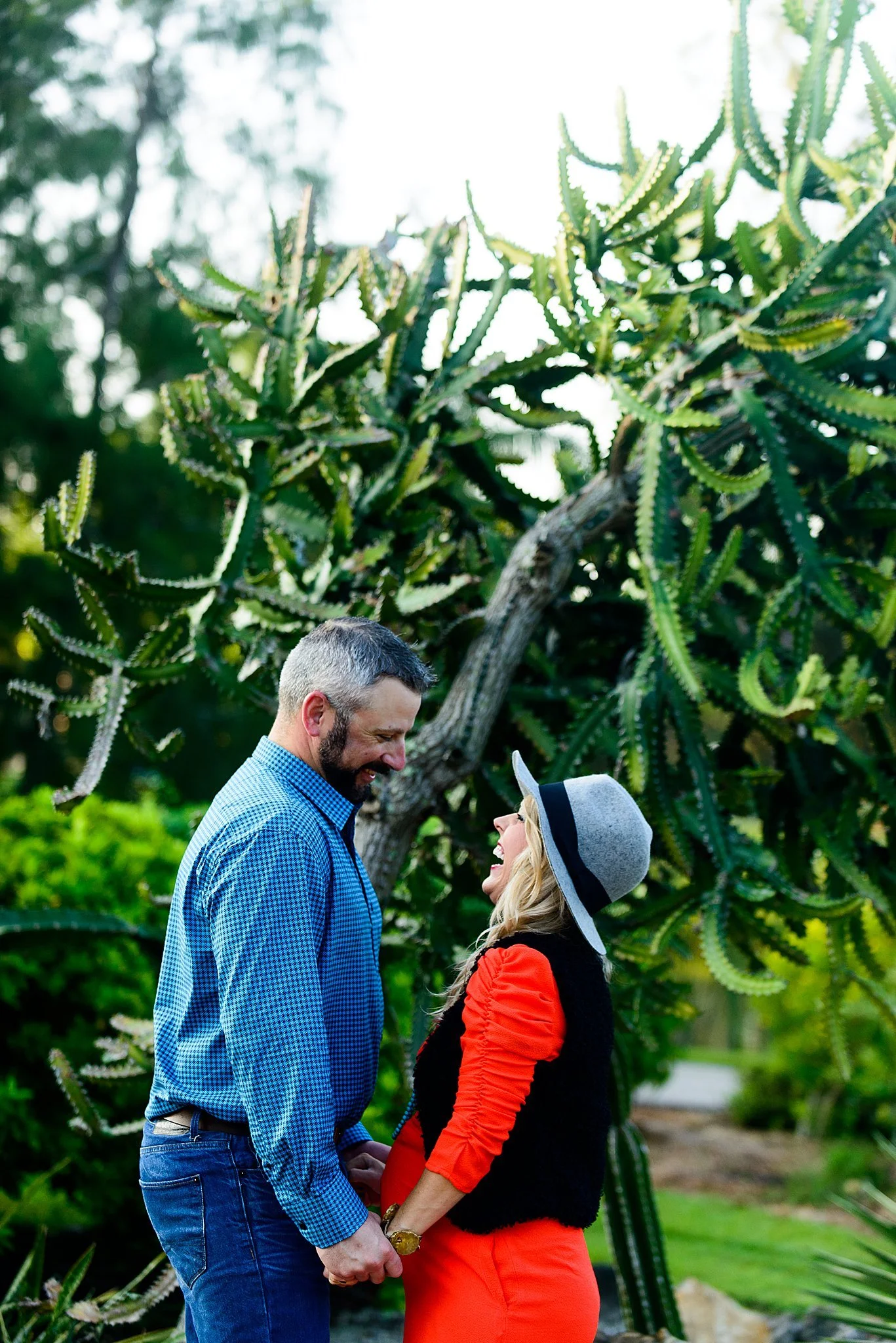 A couple holding hands and smiling at each other outdoors with a large cactus plant in the background.