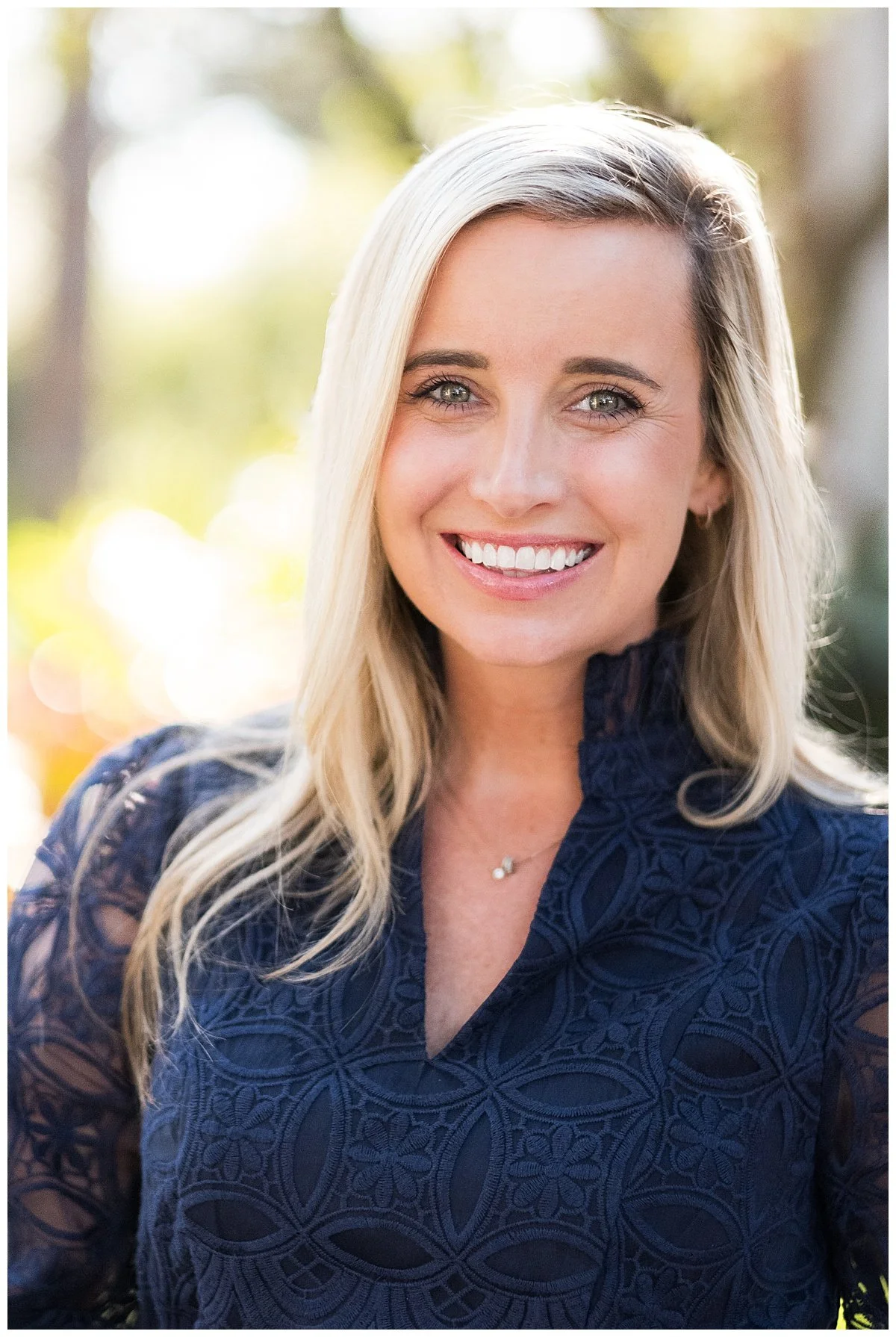 Portrait of a blonde woman smiling outdoors, wearing a dark blue lace top with a high collar and a delicate necklace, with blurred trees and sunlight in the background.