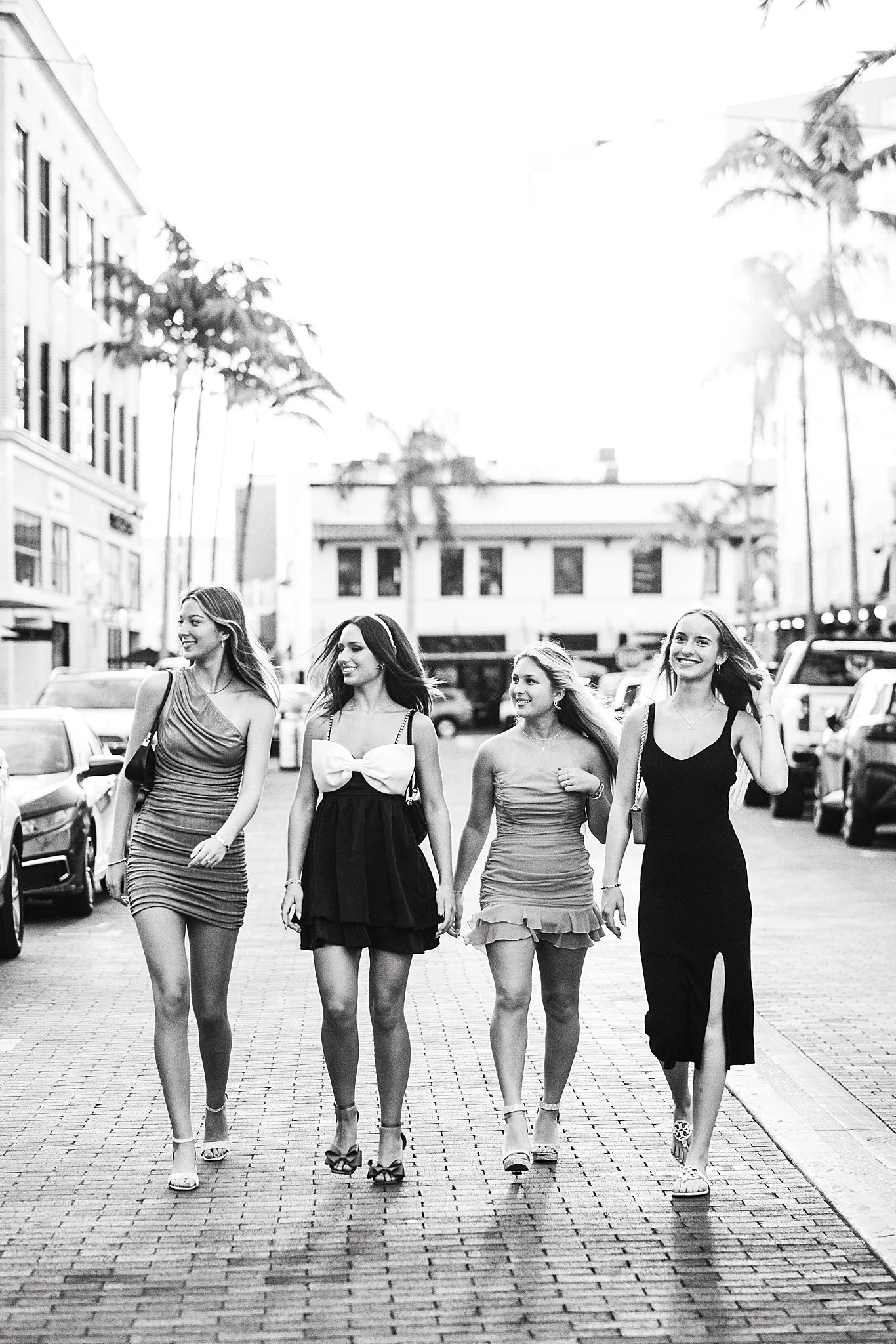 Four young women in fashionable dresses walking together on a city street, smiling, with parked cars and buildings in the background, black and white photo.