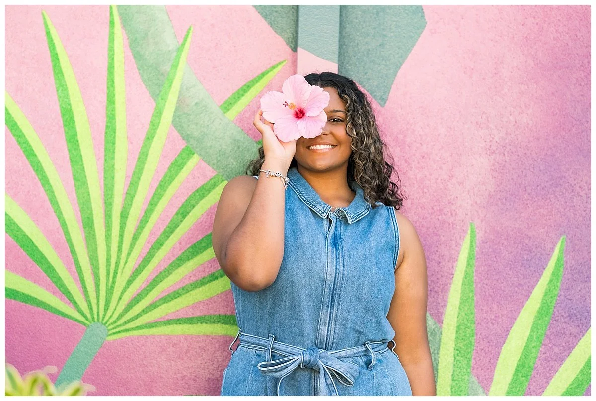A woman in a sleeveless denim dress with a tied waist holding a pink hibiscus flower in front of her face, standing in front of a colorful pink and green mural.
