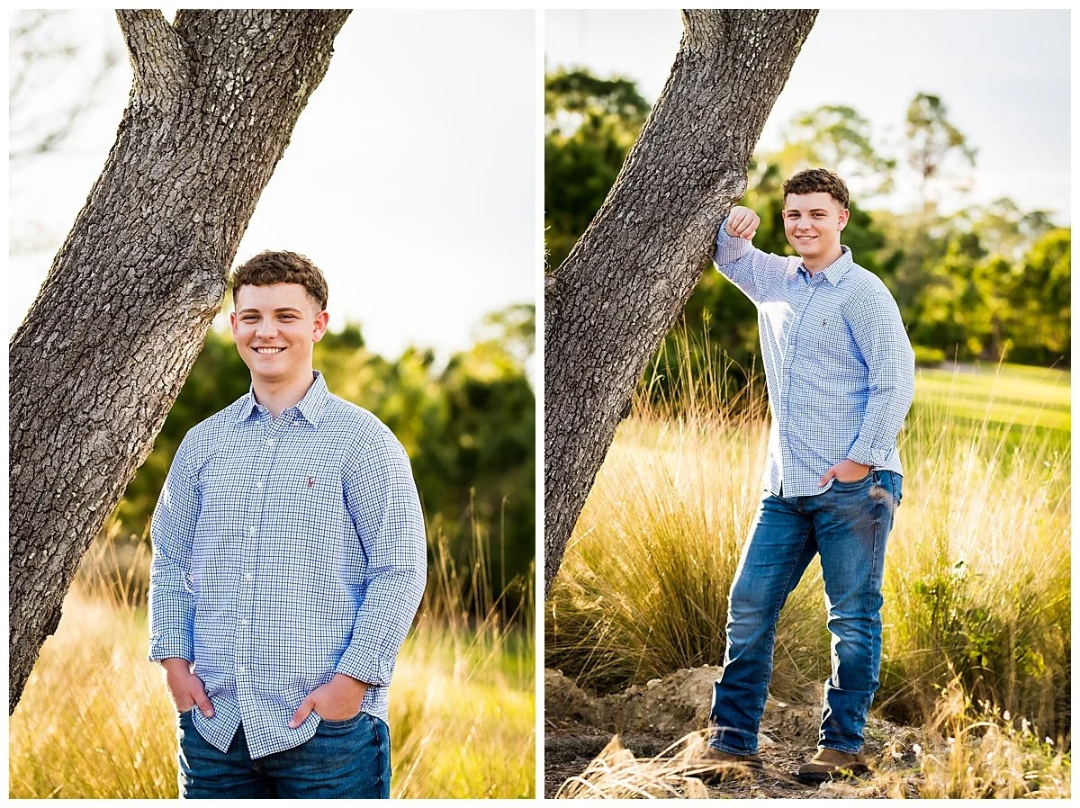A young man in a light blue checkered shirt and jeans standing outdoors next to a large tree, smiling with hands in pockets, in a grassy field with trees in the background.