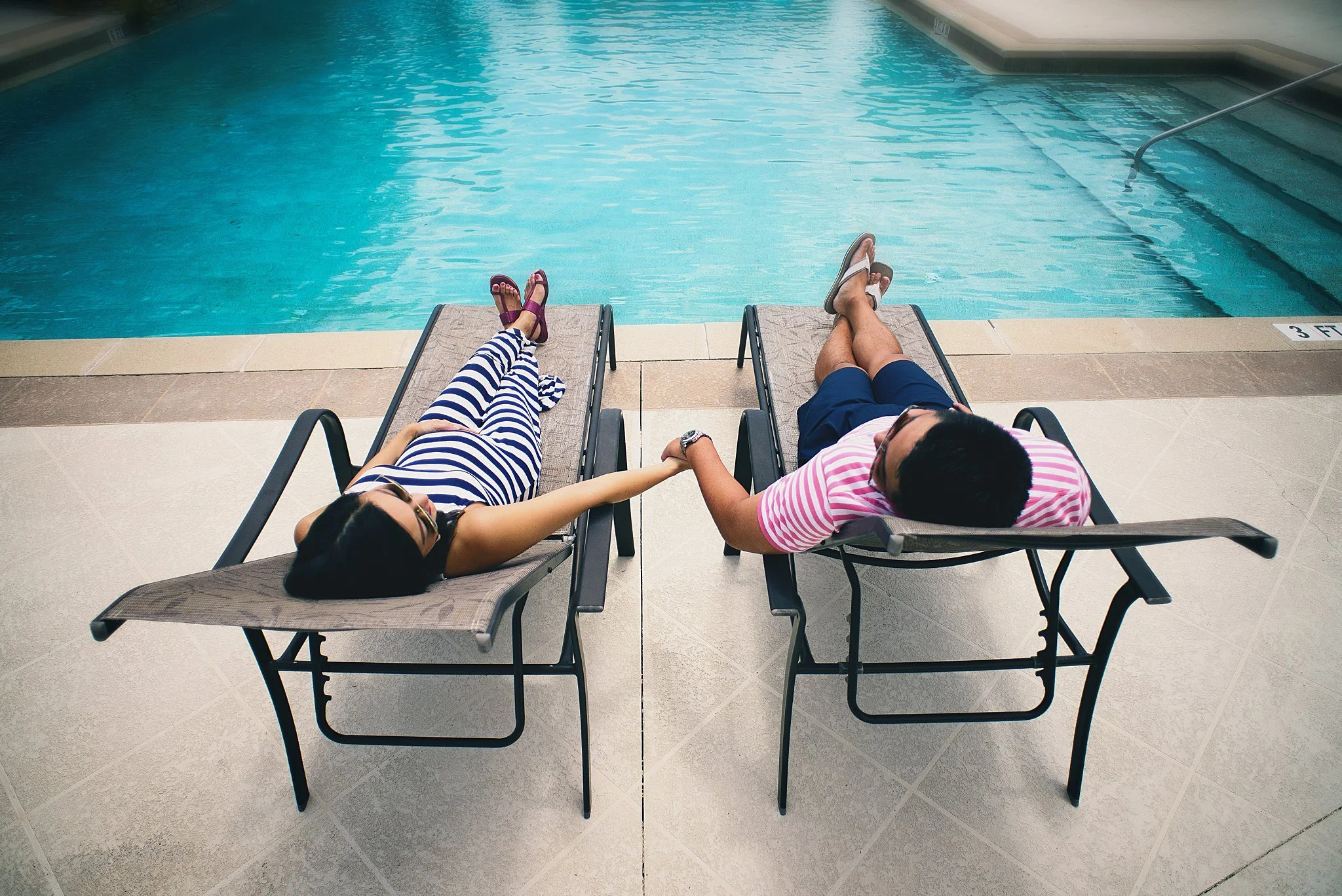 A couple is lying on lounge chairs by a swimming pool, holding hands. The woman is wearing sunglasses and a black and white striped dress, while the man is wearing a pink and white striped shirt and navy shorts.