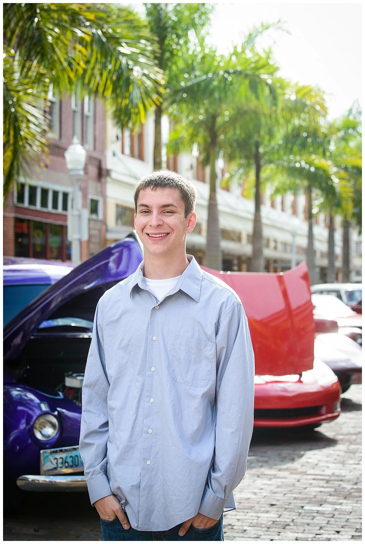 A young man smiling outdoors in front of classic cars with their hoods open, palm trees, and buildings in the background.