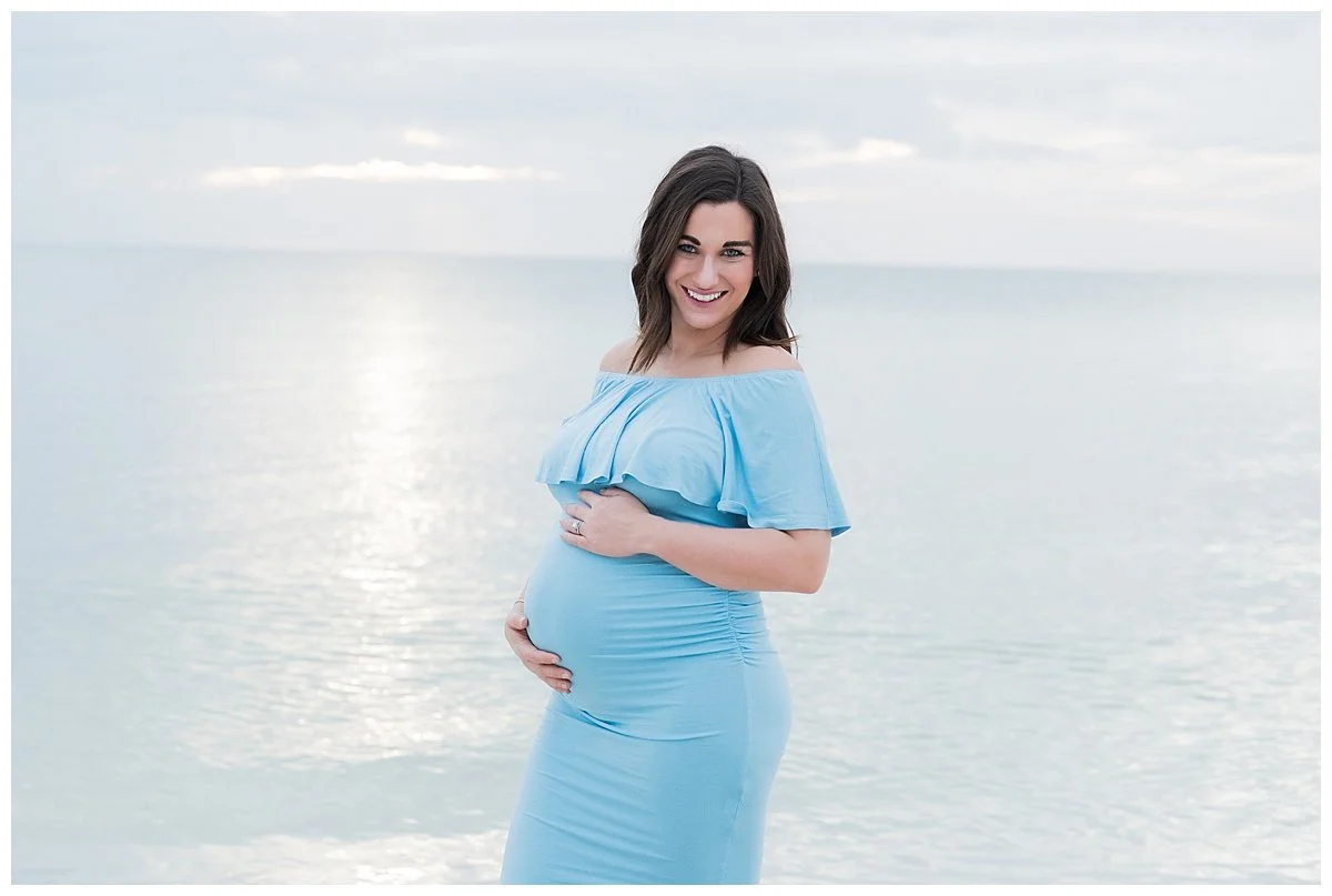 A pregnant woman in a light blue off-shoulder dress smiling at the camera, standing by a calm body of water under a cloudy sky.