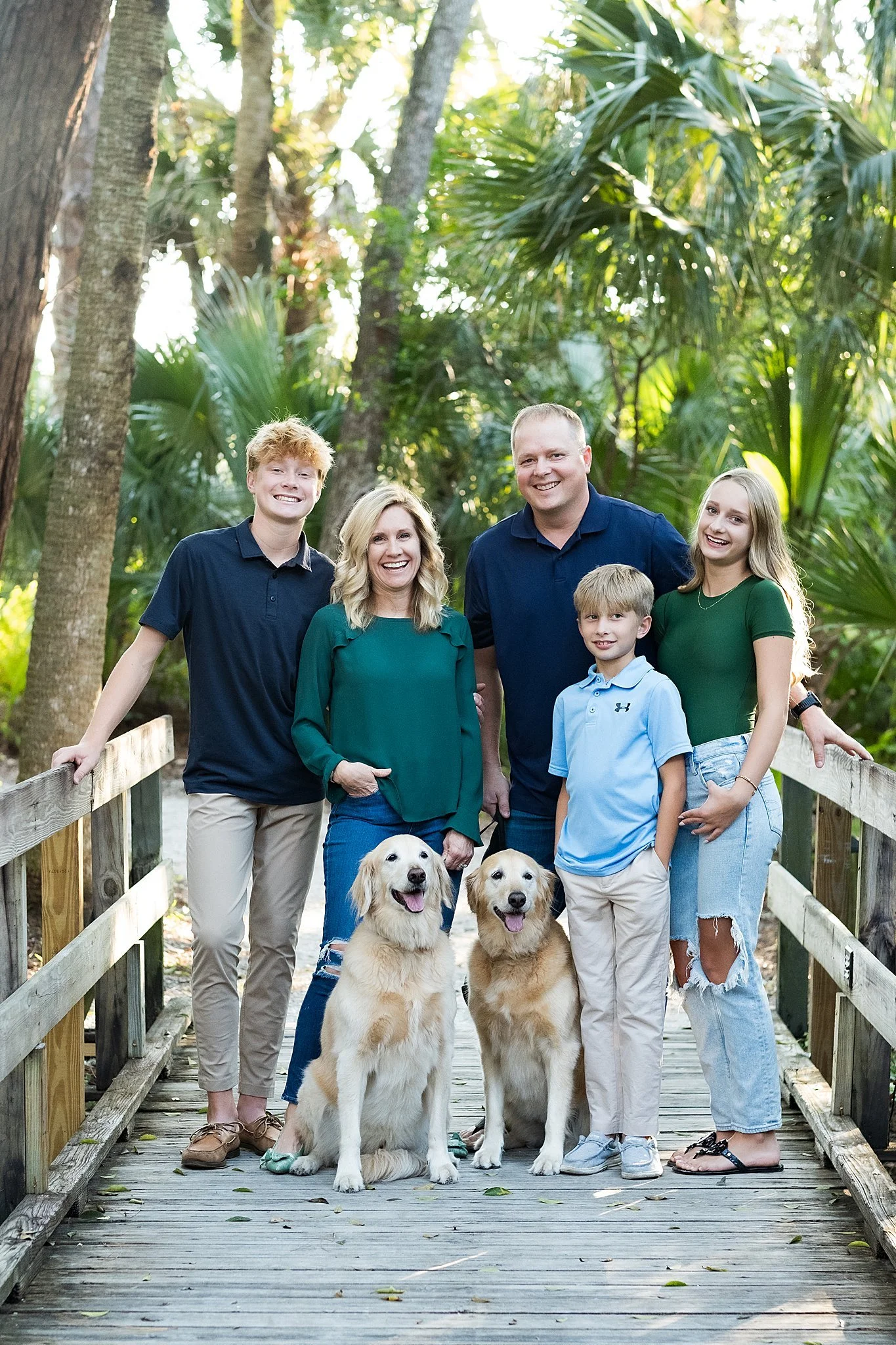 A family of six people and two dogs standing on a wooden bridge in a lush green forest, smiling at the camera.