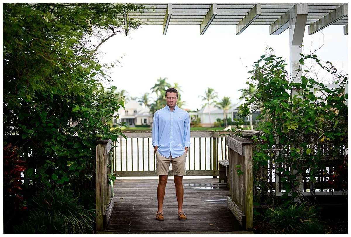 A young man standing on a wooden dock by the water, with lush greenery on either side, in a residential area with tropical trees and houses in the background.