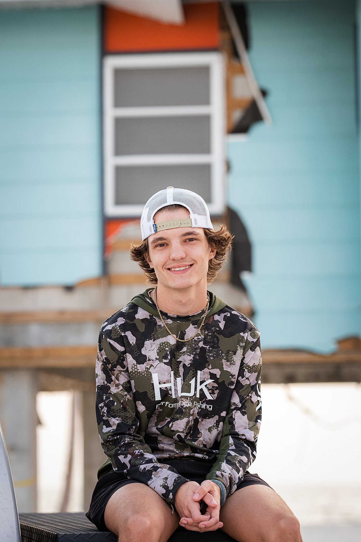 A young man sitting outdoors, wearing a camouflage hoodie, black shorts, a white and green trucker cap, and a gold chain, smiling at the camera with a colorful building in the background.