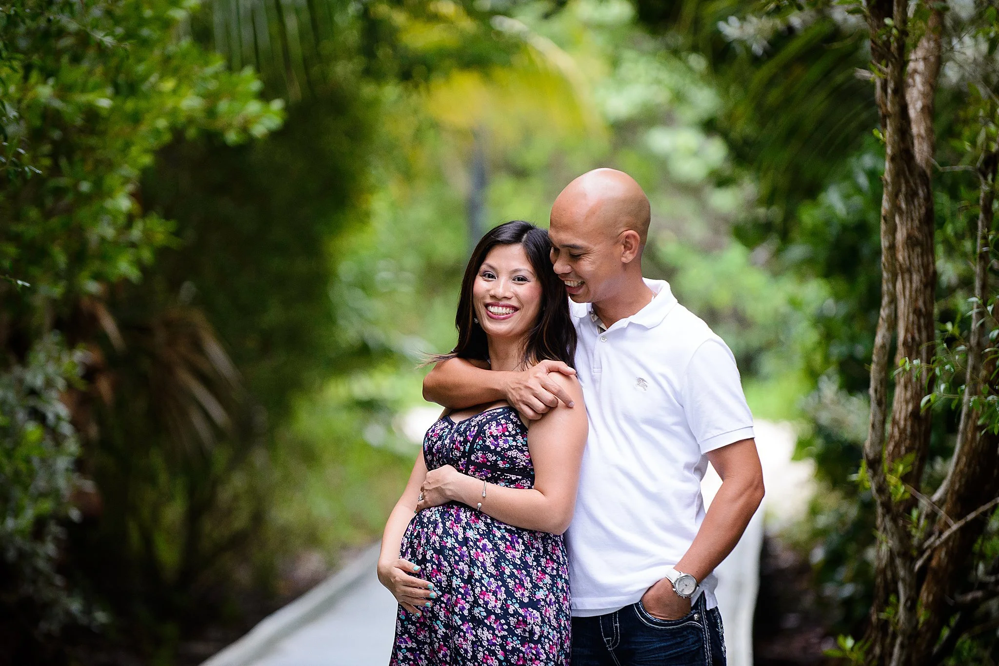 A happy couple, with the woman visibly pregnant, standing outdoors in a lush, green setting. The man is embracing her from behind, both smiling and enjoying the moment.