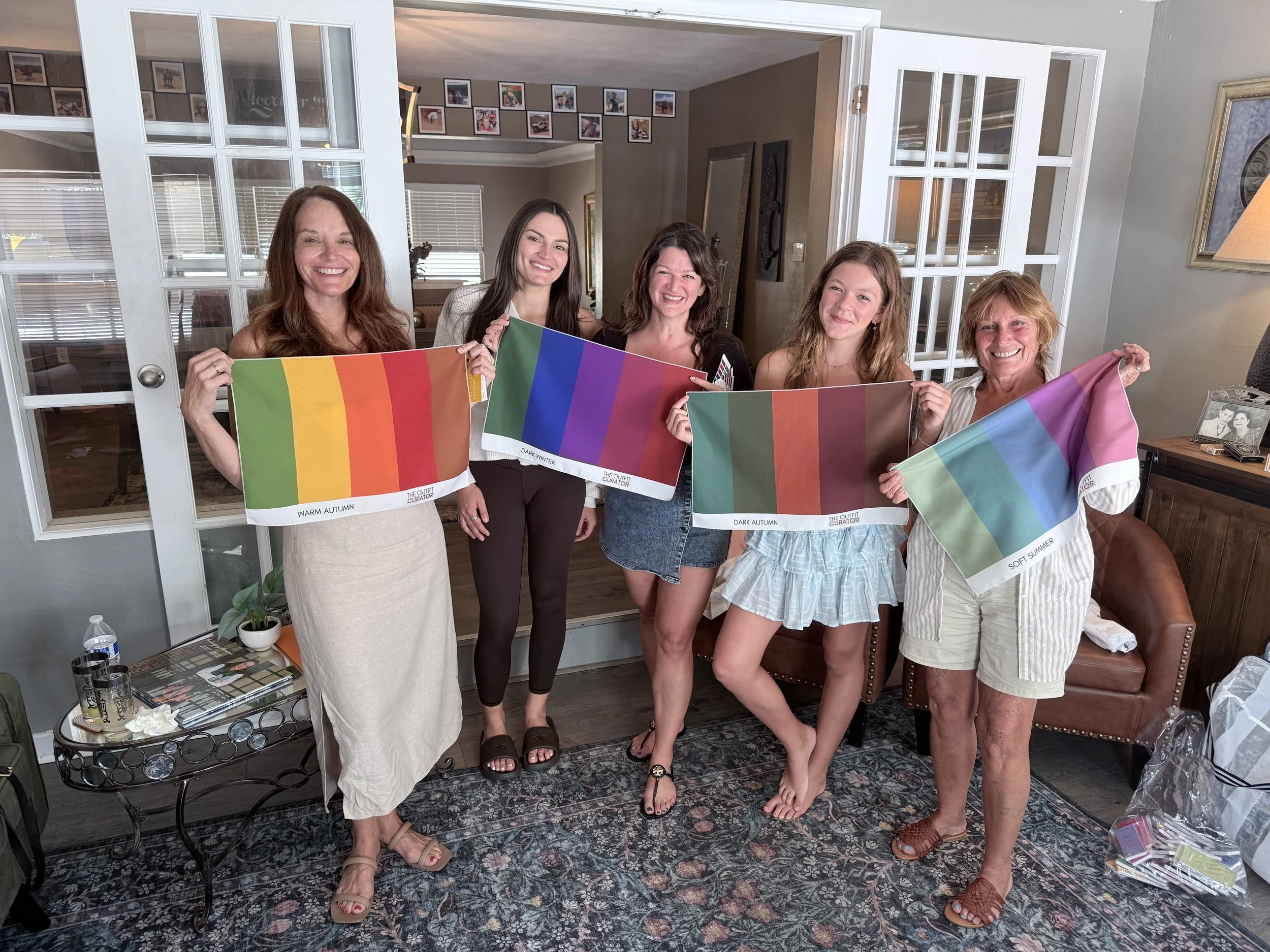 Five women standing indoors, holding rainbow-colored flags representing different color schemes, in a cozy living room.