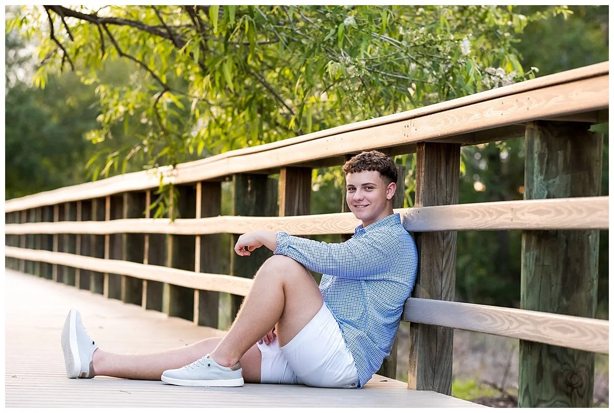 A young man with curly hair, wearing a blue checkered shirt, white shorts, and white sneakers, sitting on a wooden bridge with a lush green background, smiling at the camera.