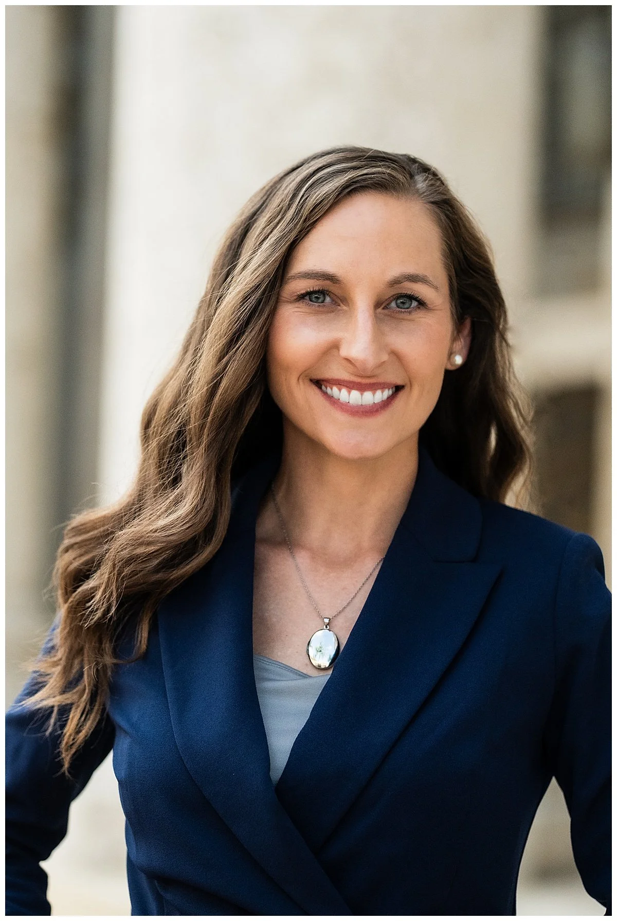 A woman with long wavy brown hair, wearing a navy blazer, a gray top, pearl earrings, and a pendant necklace, smiling outdoors.