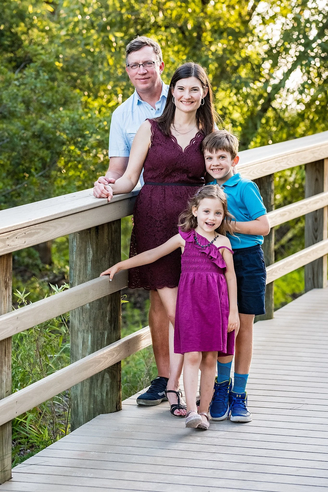 A family of four standing on a wooden bridge outdoors surrounded by trees, with a man, a woman, a young boy, and a young girl smiling at the camera.