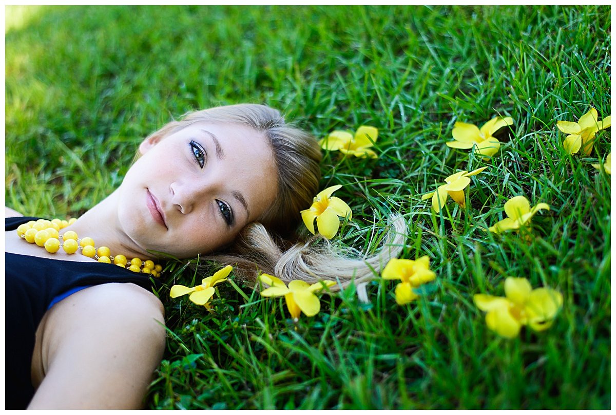 A woman with blonde hair lying on green grass surrounded by yellow flowers, wearing a black top and a yellow beaded necklace.