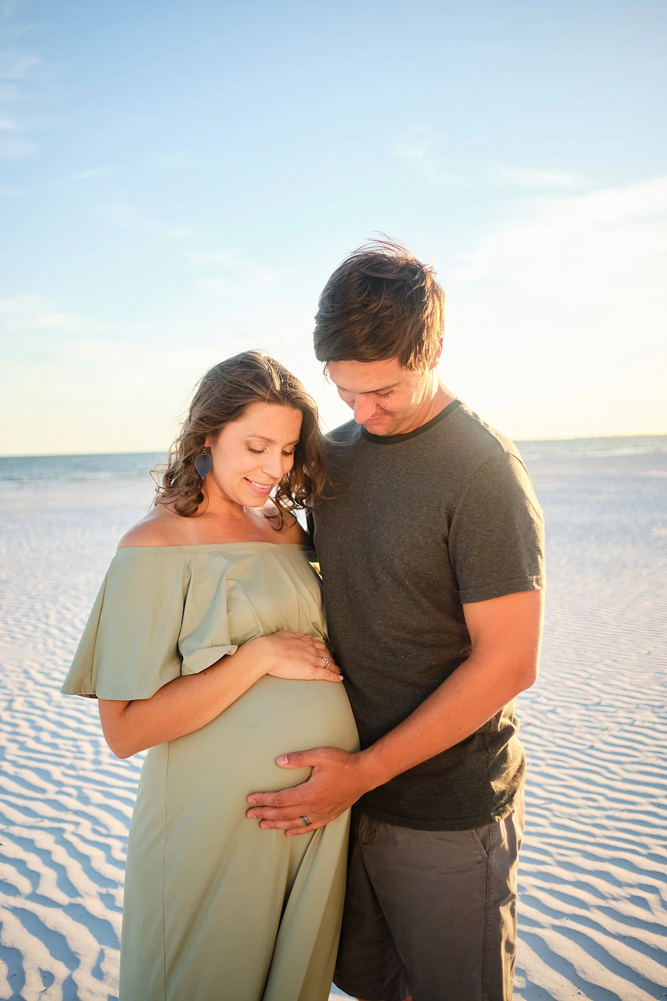 A pregnant woman and a man embracing on a beach, both touching her belly and looking down at it.