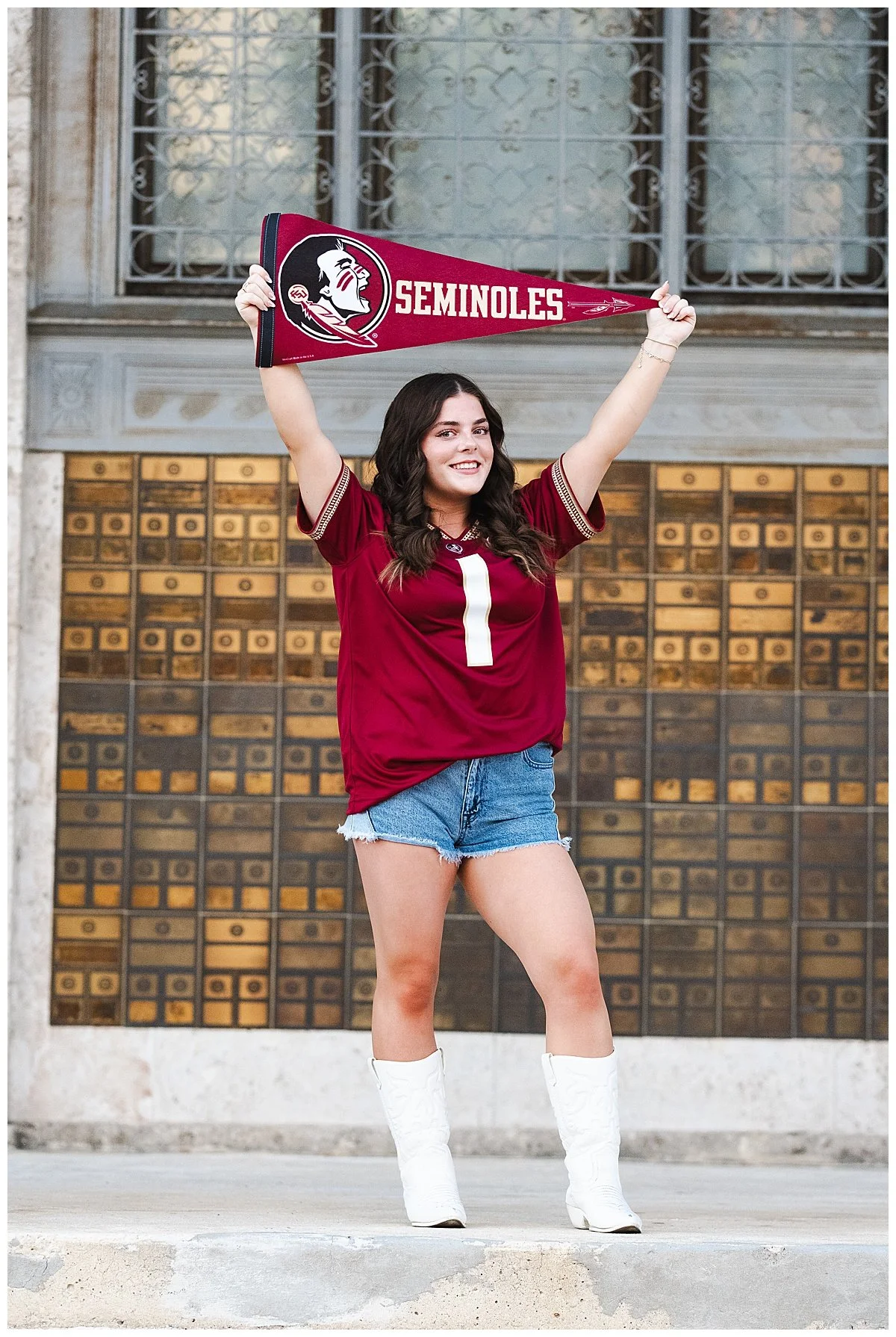 Young woman in a Florida State Seminoles football jersey and white cowboy boots holding a Seminoles pennant, standing in front of a decorative wall.