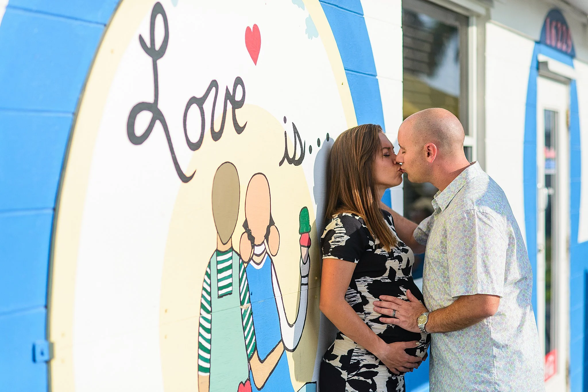 A couple kissing in front of a colorful wall with a mural of women holding ice cream cones and the word "love" written on it.