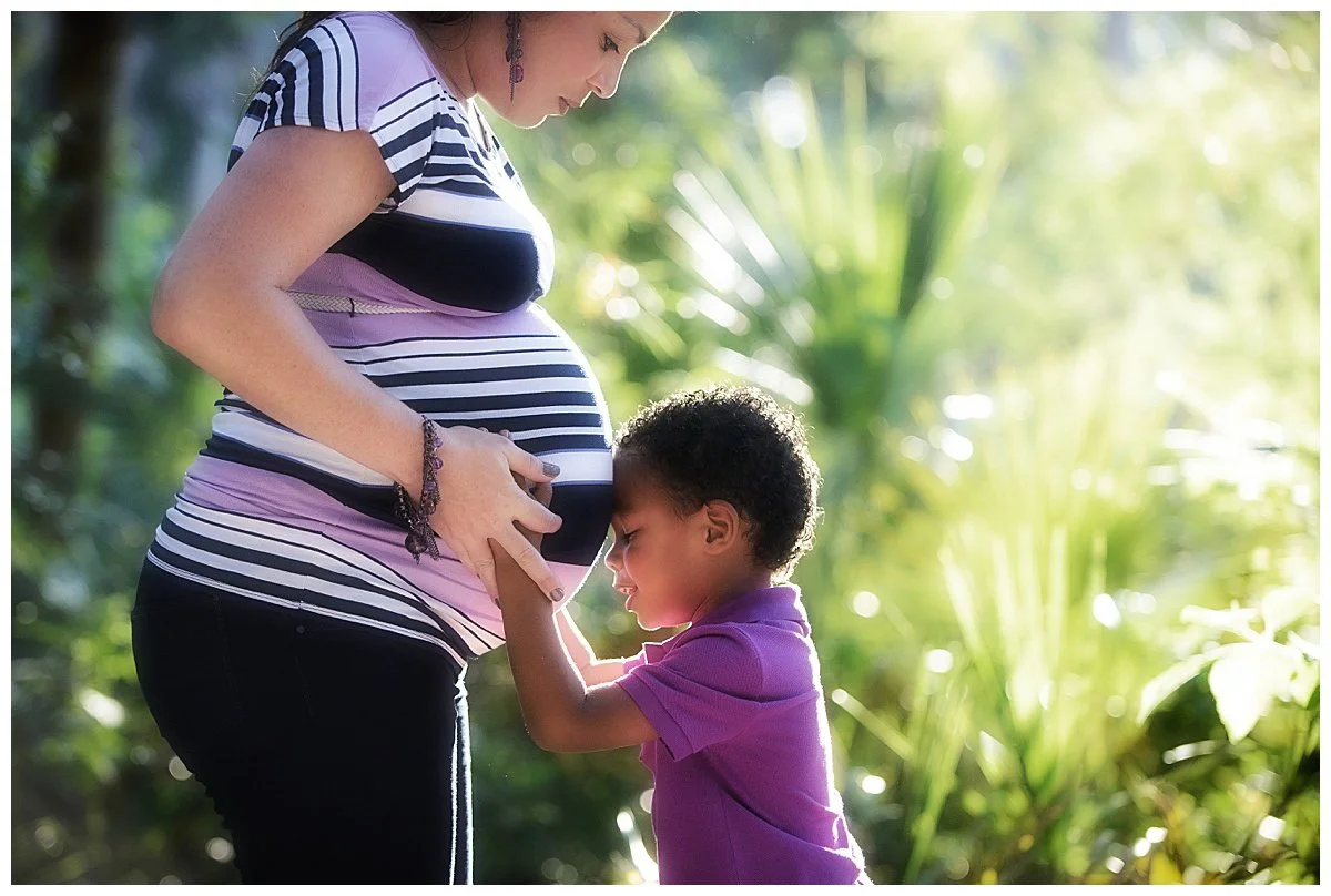 A pregnant woman and a young boy with curly hair standing outdoors in a lush, green garden. The boy is touching the woman's belly with his eyes closed, and she is looking down at him. The sunlight filters through the trees, creating a warm and peacef