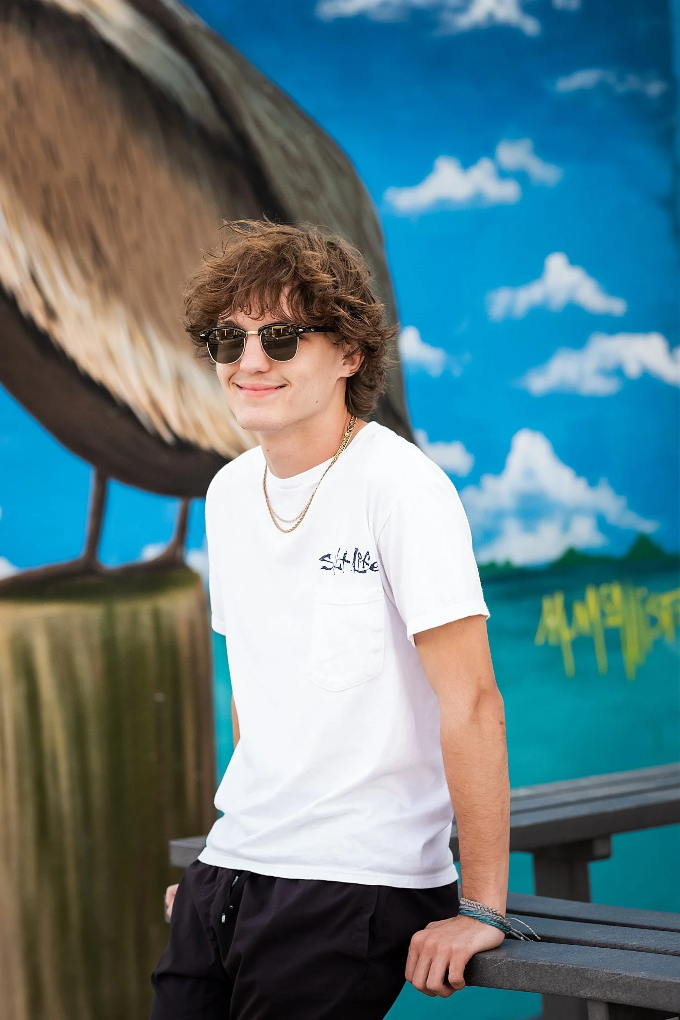 A smiling young man with curly brown hair wearing sunglasses, a white t-shirt, and black pants, standing outdoors near a railing.