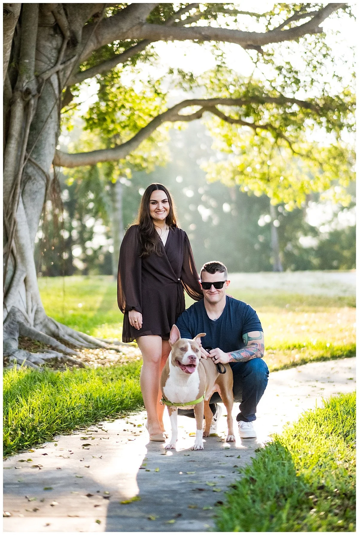 A woman and a man with a dog standing under a large tree on a park path during sunlight.