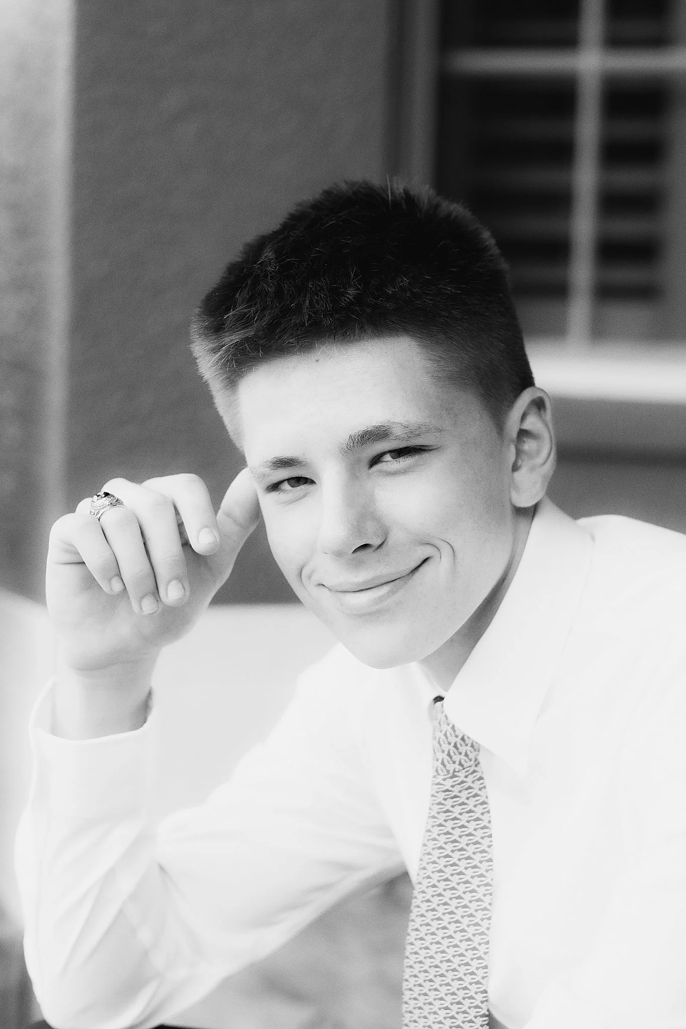 A young man smiling, wearing a white shirt and patterned tie, resting his chin on his hand in a professional setting.