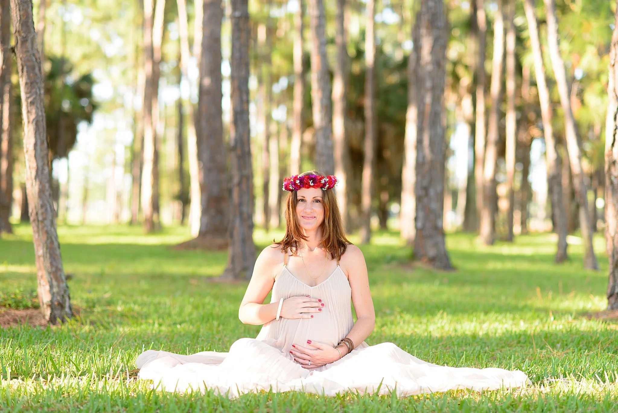A pregnant woman with red hair and a floral headband sitting on grass in a forest, wearing a flowing white dress, smiling, with her hands on her belly.