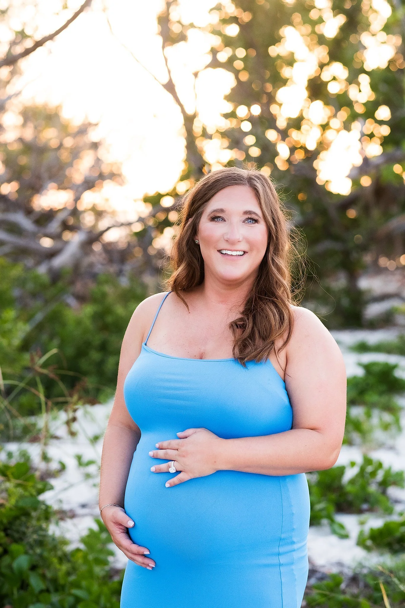 A woman in a blue dress stands outdoors near trees with sunlight in the background, smiling and holding her pregnant belly.