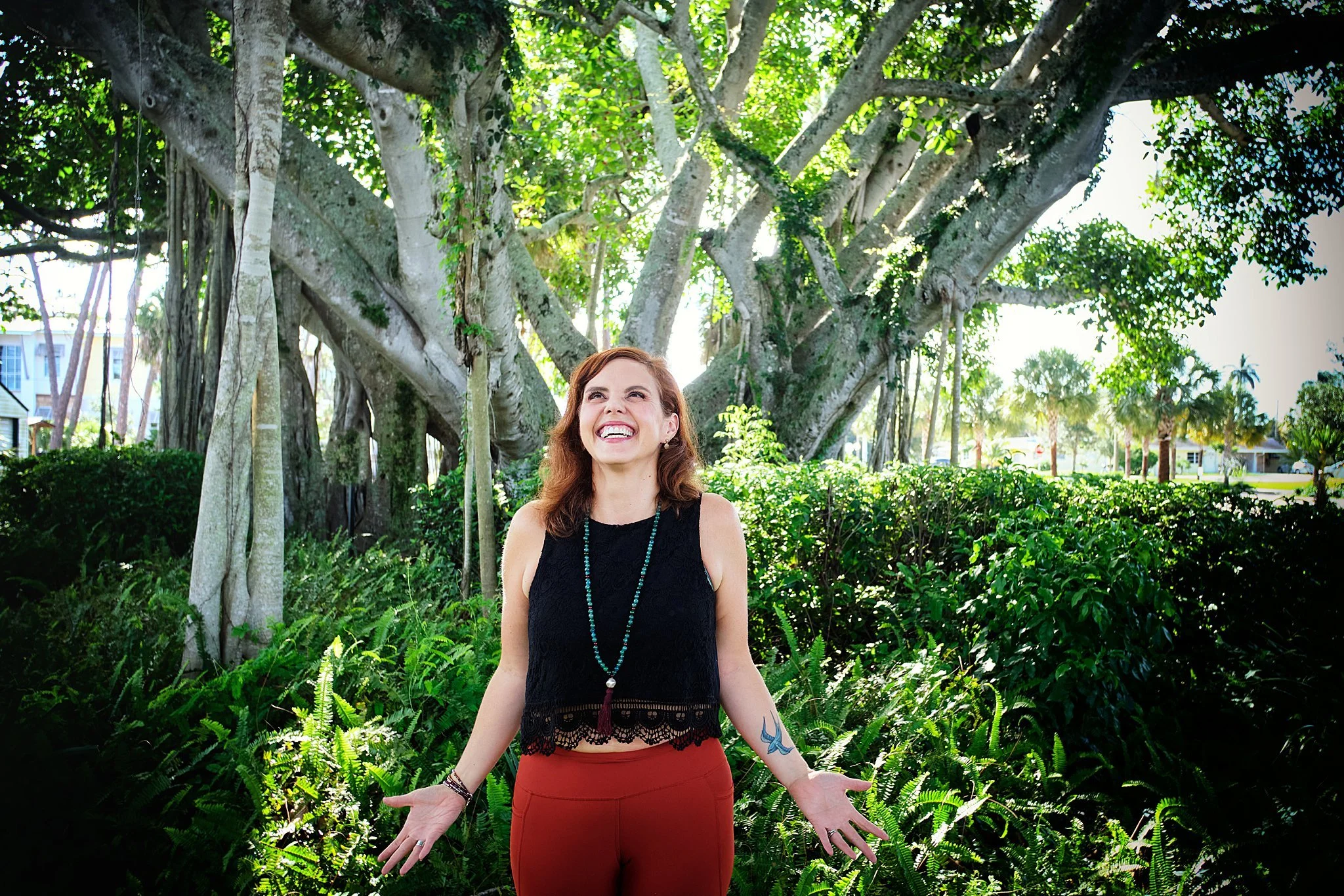 A woman with brown hair smiling and looking up outdoors near a large tree with lush green foliage.
