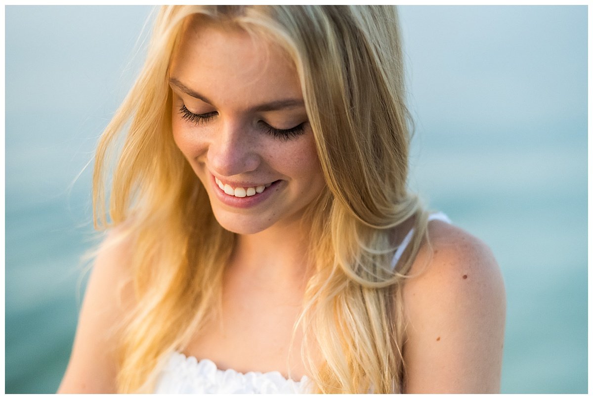 A young woman with blonde hair and freckles smiling with her eyes closed, standing outdoors near water.