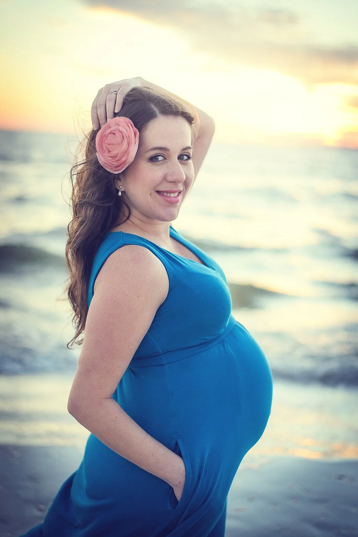 A pregnant woman wearing a blue dress, with a pink flower in her hair, smiling and posing on the beach at sunset.