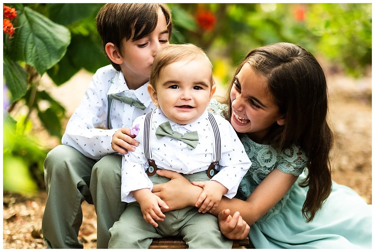 A woman with long brown hair smiling and playing with a toddler boy, while an older boy kisses the toddler's head, outdoors surrounded by green plants and red flowers.