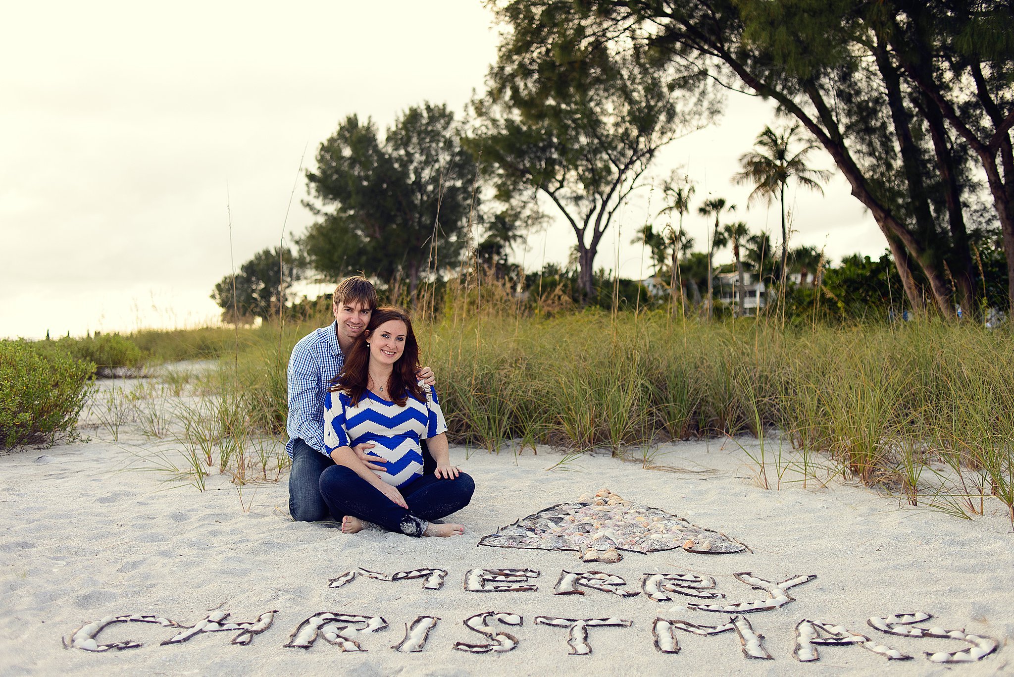 A couple sitting on the beach with the words 'Papa Chris's baby' spelled out in shells on the sand in front of them