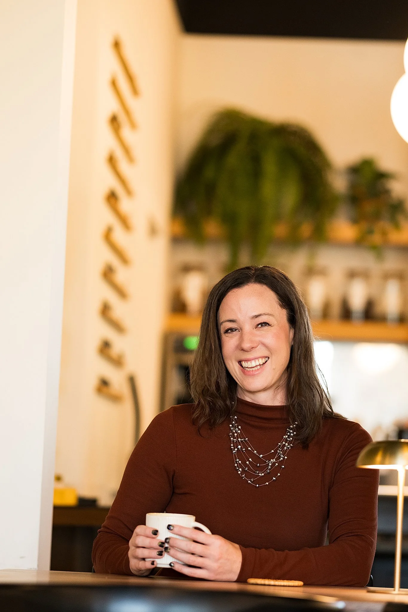 A woman smiling and holding a mug in a cozy cafe or coffee shop, with warm lighting and indoor plants in the background.