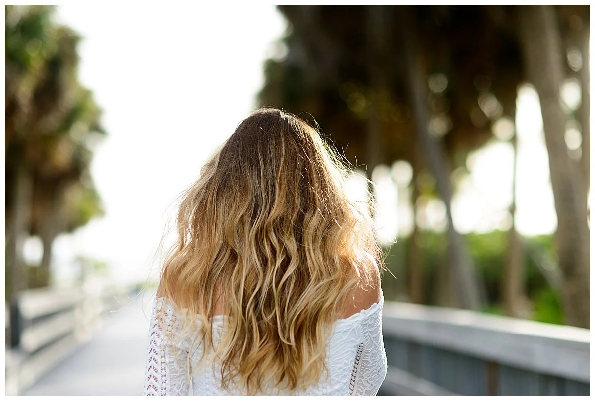 Back view of a woman with long, wavy blonde hair walking on a bridge surrounded by trees.