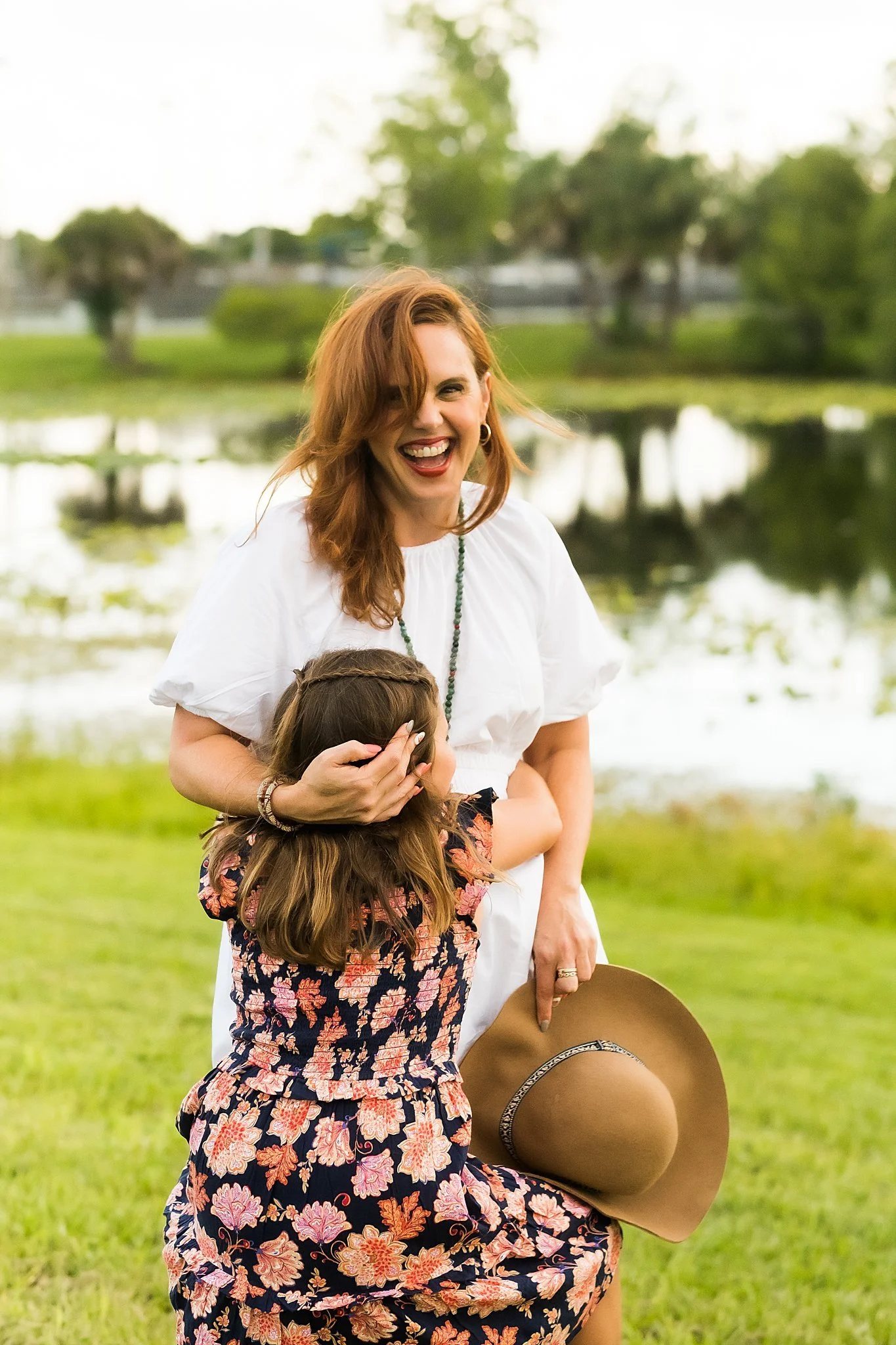 A woman with red hair smiling and laughing while hugging a young girl outdoors near a pond with green trees in the background.