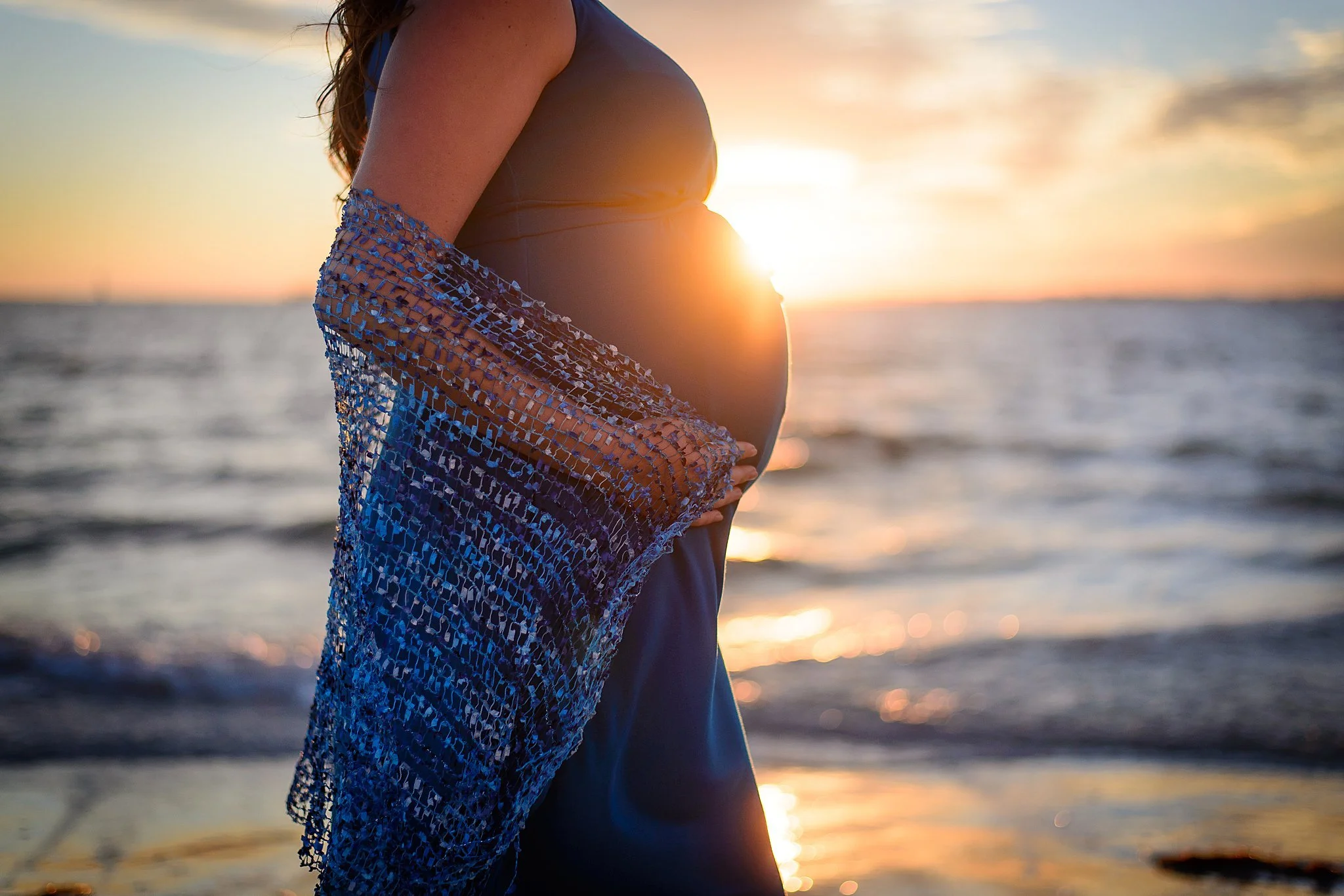 Pregnant woman at the beach during sunset, holding her belly and draped in a blue shawl.