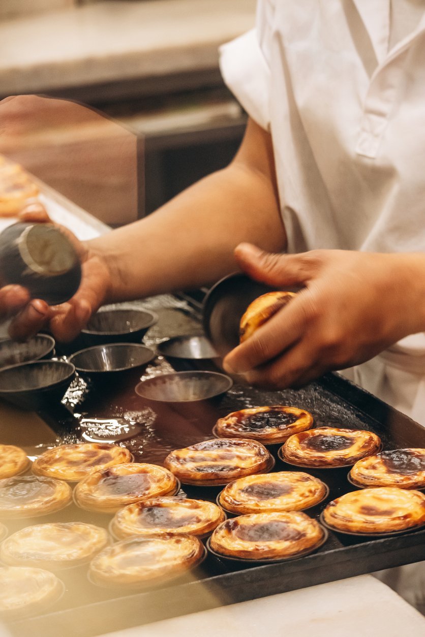 Person preparing traditional Portuguese custard tarts, known as Pastéis de Nata, on a baking tray in a bakery kitchen.