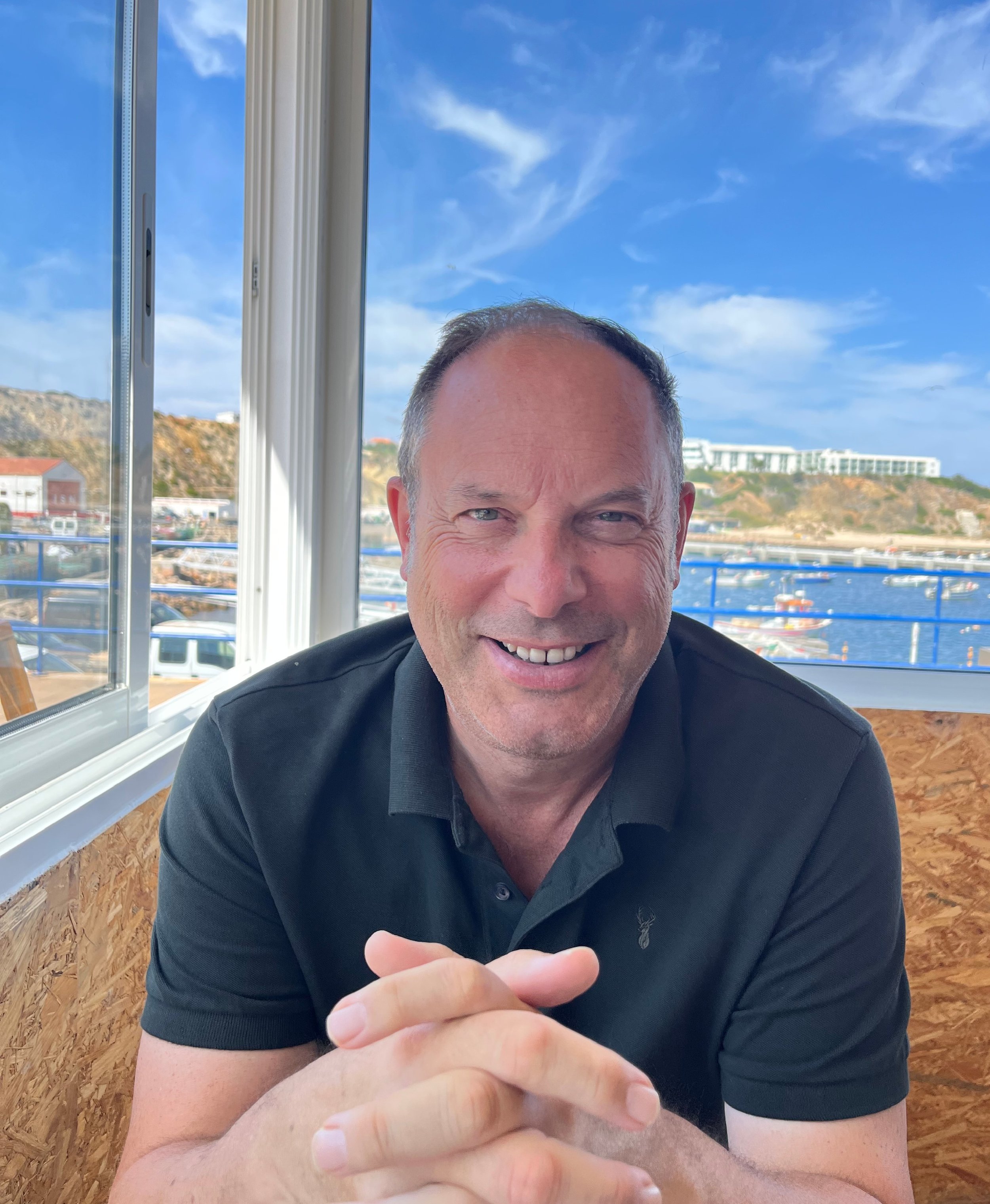 A smiling man with short hair sitting at a table near a window with a view of boats and a hillside harbor.