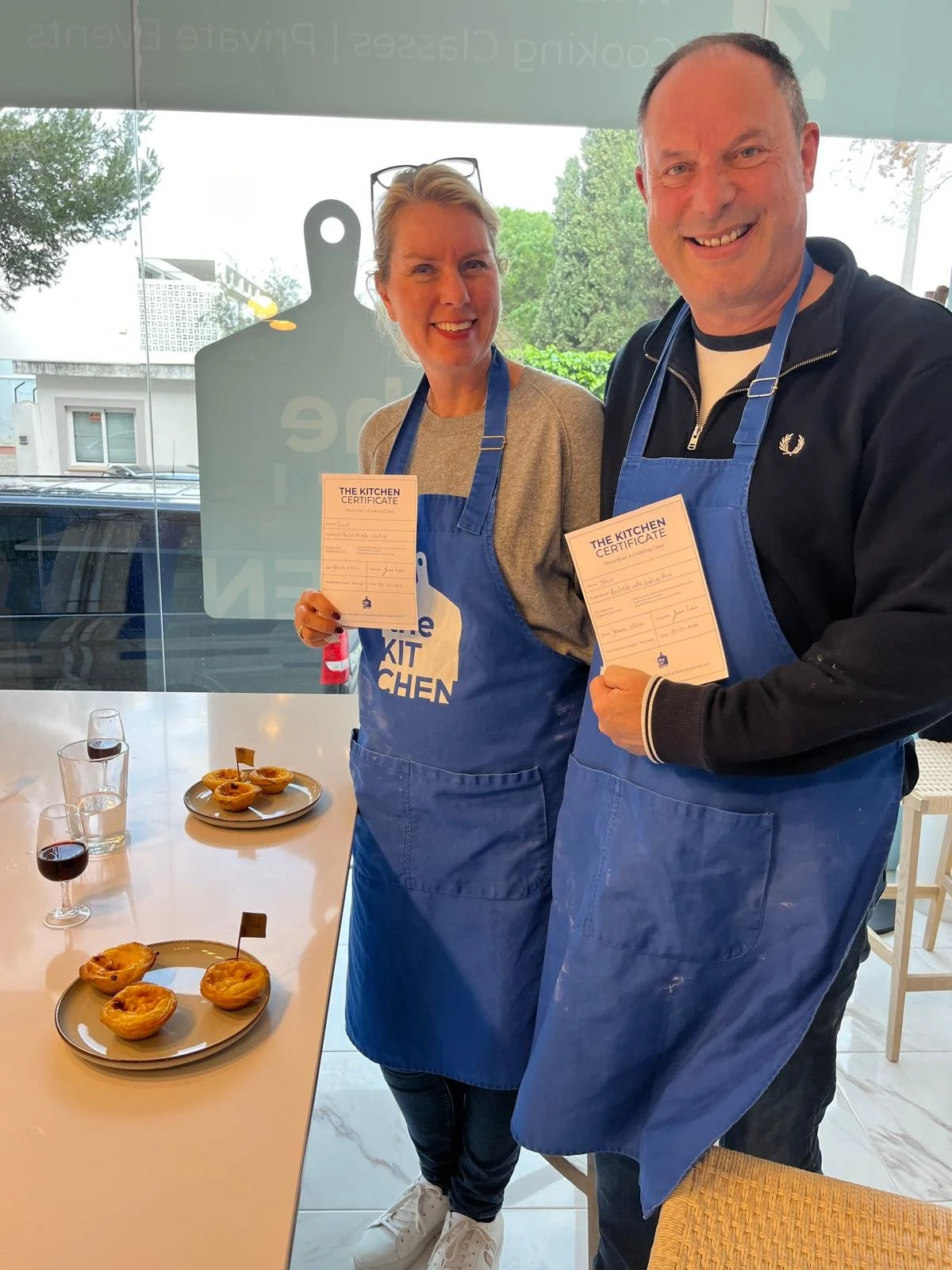 Two smiling people standing in a pastel de nata cooking class, each holding a certificate. They are wearing blue aprons with a white logo. In front of them on a white table are pastel de natas and glasses of port.