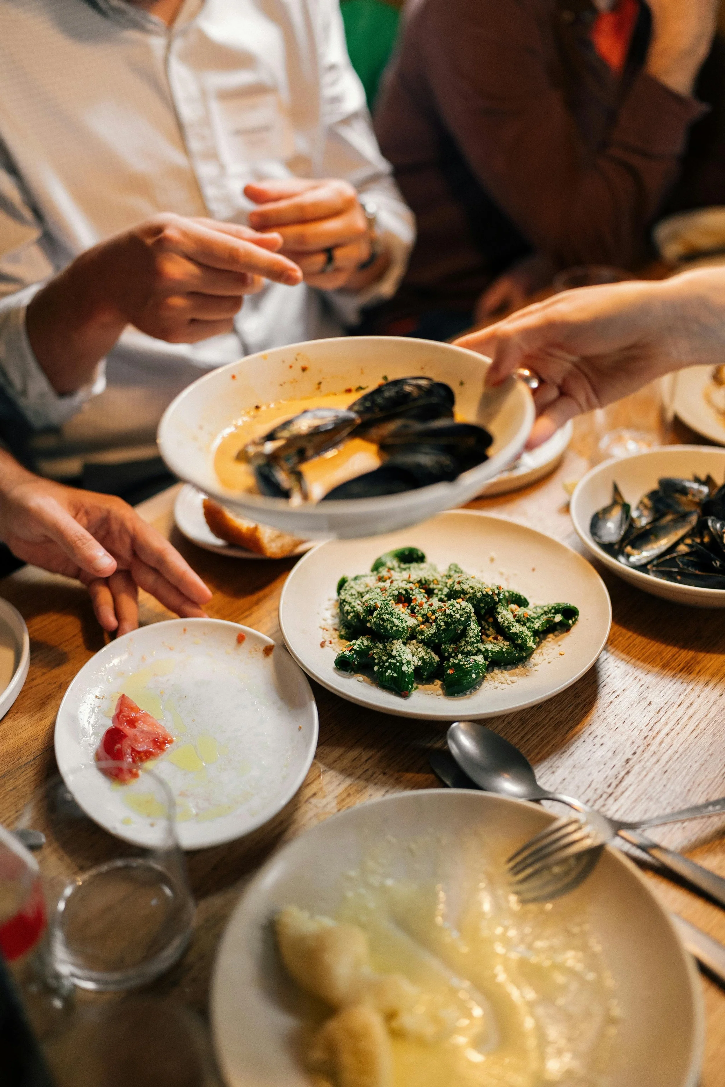 People sharing a meal with plates of mussels, pasta, and salad on a wooden table.