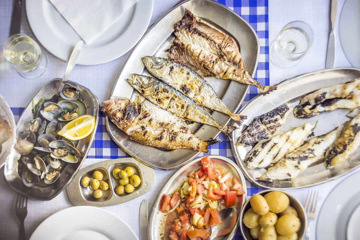 Assorted grilled fish dishes on a dining table with sides of vegetables, olives, and clams.