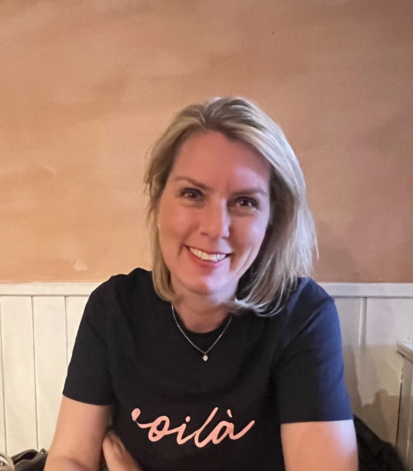 A woman with shoulder-length blonde hair smiling, wearing a black T-shirt with pink cursive text, sitting in a room with beige walls.