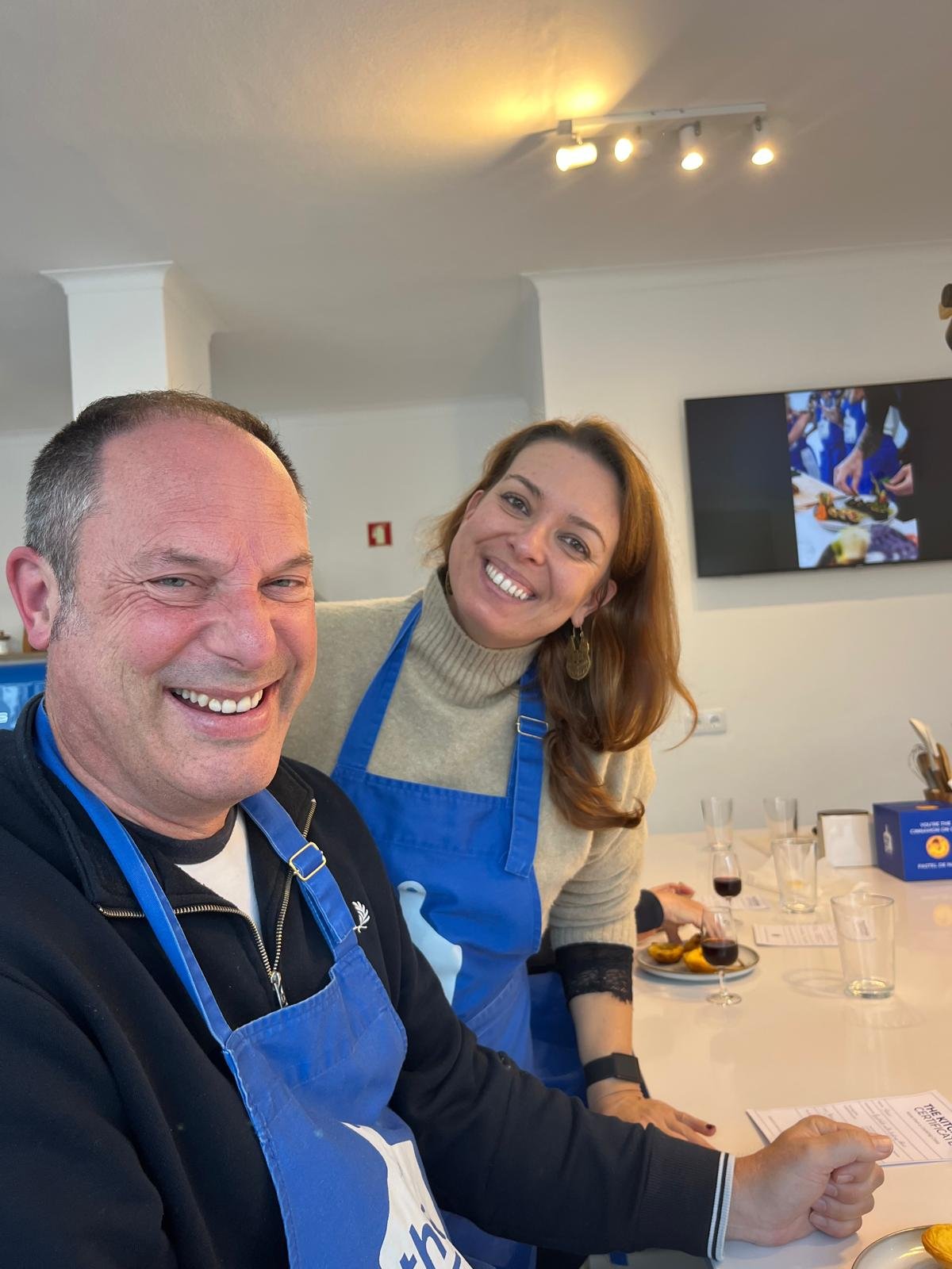 A man and a woman smiling at a pastel de nata cooking class with glasses and plates, wearing blue aprons in a brightly lit room.