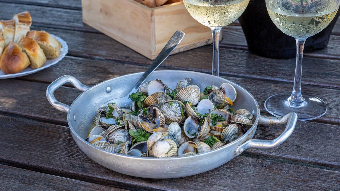 A metal pan filled with cooked clams garnished with herbs sits on a wooden table, with two glasses of white wine and a plate of bread rolls nearby.